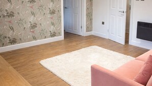 A living room with a wooden floor, a white rug, a pink sofa, and floral wallpaper. - Spacious 2-BR Annexe near Abington Park - 10% off (West Northamptonshire)