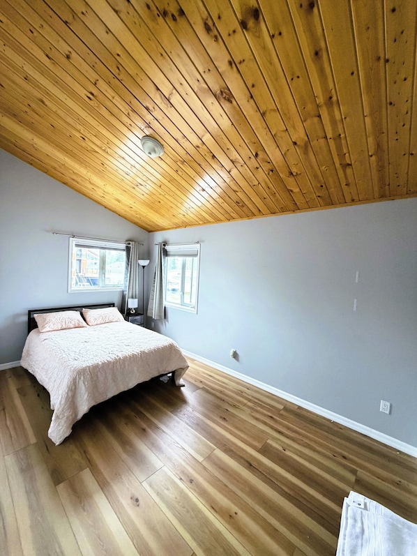 A bedroom with a wooden ceiling, a bed with a white bedspread, two windows with curtains, and a wooden floor.