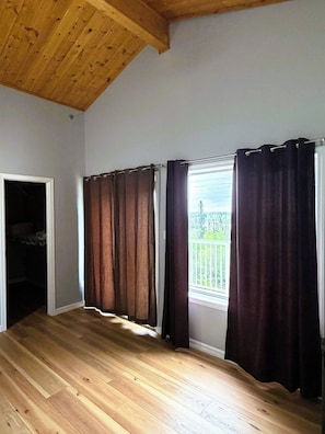 A room with wooden flooring, a window with curtains, and a visible balcony.