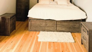 A bedroom with a wooden ceiling, a bed with white bedding, a nightstand, a dresser, and a white rug.