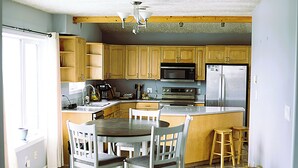 A kitchen with wooden cabinets, a microwave, a refrigerator, a dining table with chairs, and a small stool.