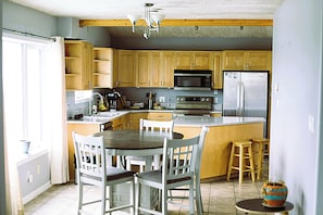 A kitchen with wooden cabinets, a microwave, a refrigerator, a dining table with chairs, and a small stool.