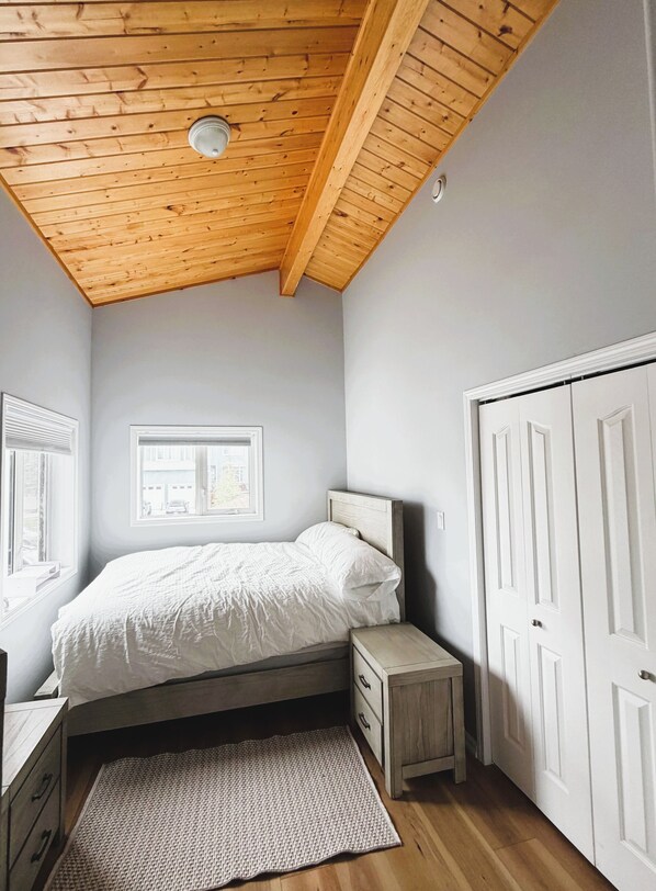 Une chambre avec un plafond en pente, un lit, une fenêtre, une porte et une petite table de chevet.