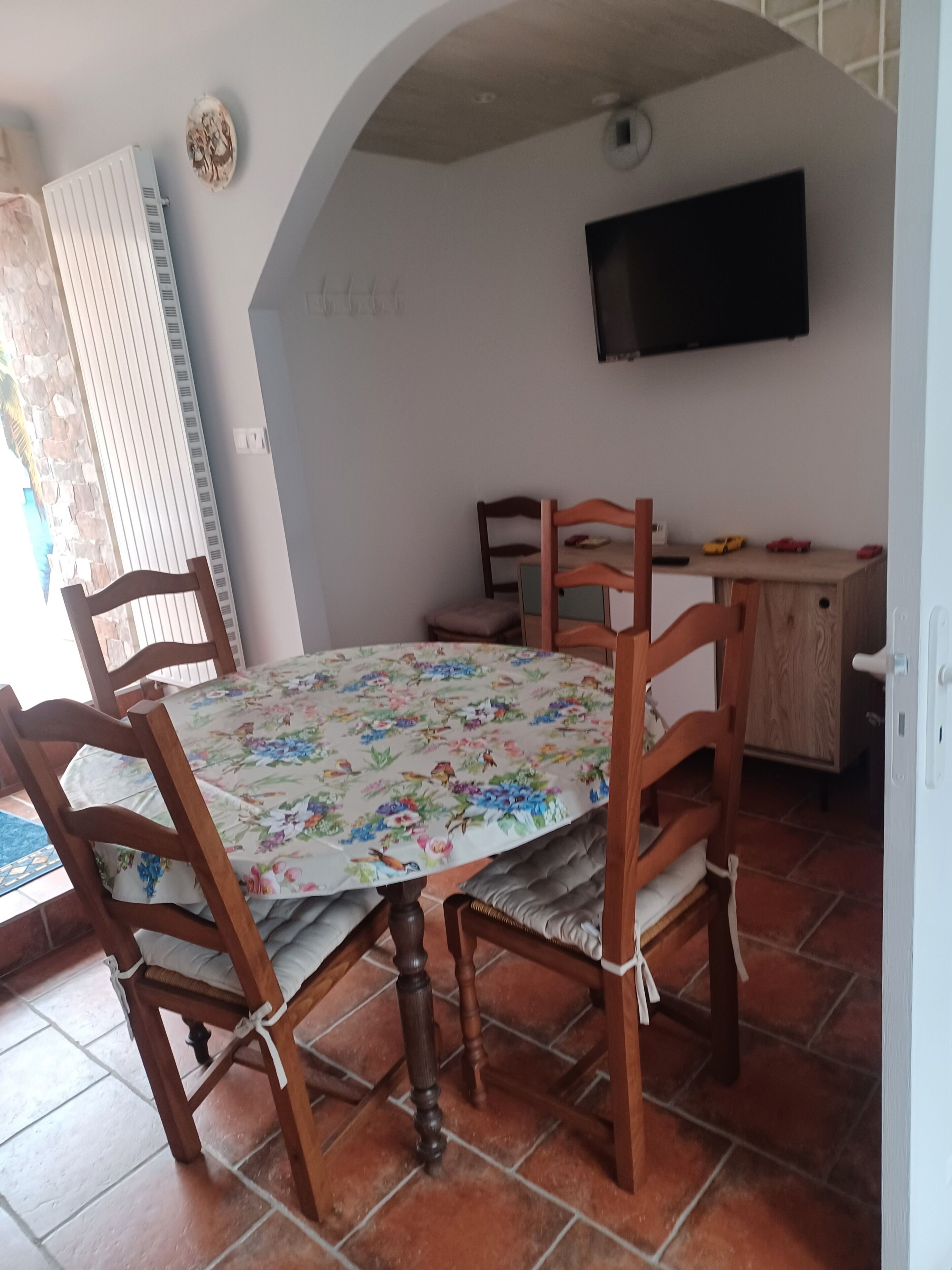 Ein Essbereich mit einem Tisch, gedeckt für vier Personen, umgeben von Holzsesseln. Über dem Tisch ist ein Fernseher an der Wand montiert.