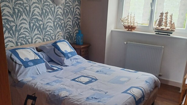 A bedroom with a bed, a nightstand, a window with a view of ships, and a patterned wallpaper. - Les Orangers Vacation Rental (Pleubian)