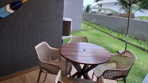 A patio with a round table and wicker chairs, a wall-mounted dartboard, and a view of a grassy area with a palm tree. - F.206 | Your Refuge with comfort and good taste (Tamandaré)