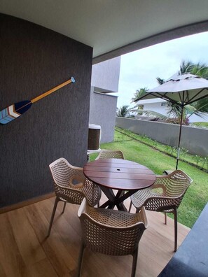 Eine Terrasse mit rundem Tisch und Korbstühlen, einem an der Wand befestigten Dartspiel und Blick auf eine Wiese mit einer Palme.