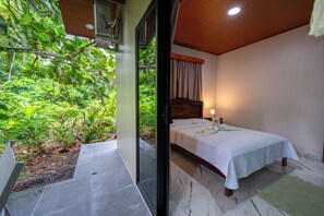 Ein Schlafzimmer mit Bett, Nachttisch, Lampe und Blick auf Grün hinter einer Glasdoor.