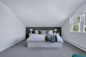 A bedroom with a large bed, two bedside tables with lamps, a window with a view of trees, and a grey carpet.