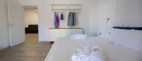 A hotel room with a bed, a bedside table, a bench with hanging clothes, and a door leading to another room.