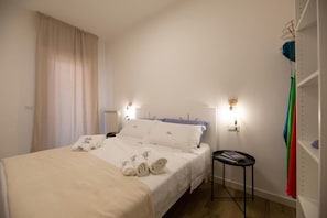 Ein Schlafzimmer mit einem Bett, zwei Nachttischlampen, einem Nachttisch, einem kleinen Tisch und einem Regal mit gefalteter Kleidung.