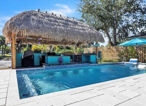 A swimming pool with a thatched roof structure, a bar area, and a wooden fence.