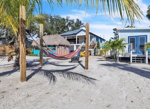 Een strandlandschap met een hangmat, een constructie met rieten dak en een blauw gebouw.