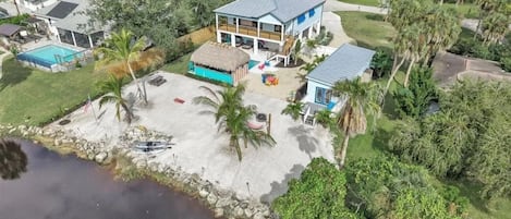 Aerial view of the waterfront residence with a sandy backyard, tiki bar, and outbuilding.