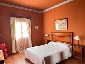 Un dormitori amb un llit, dues taules de nit, una petita taula i una finestra amb cortines lleugeres, i un quadre al mur.