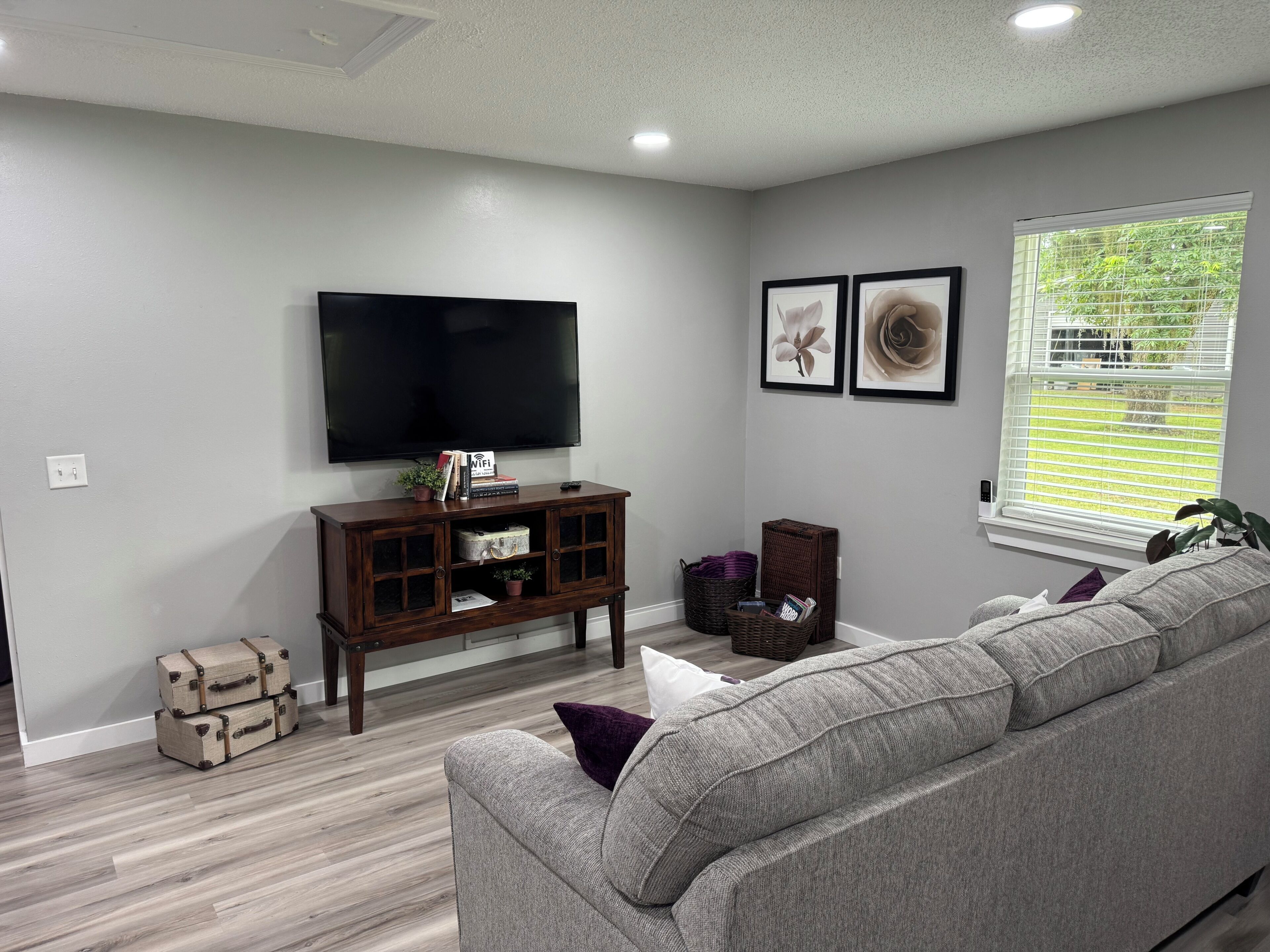 A living room with a flat-screen TV, a grey sofa, a wooden cabinet, and a window with blinds.