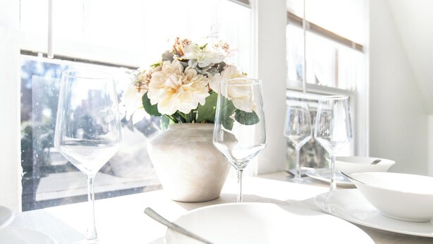 Et spisebord dækket med hvide tallerkener, en skål og en vase med blomster.