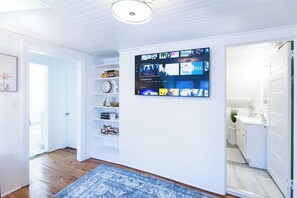 A hallway with a mounted TV displaying various channels, a blue patterned rug, and a white door leading to a bathroom. - 1 Block Off Main Downtown Greenville (Greenville)
