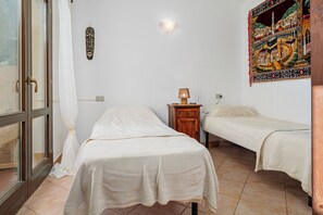 Ein Zimmer mit zwei Betten, einem Nachttisch aus Holz mit einer Lampe, einer Wandleuchte und einem farbenfrohen Wandteppich.