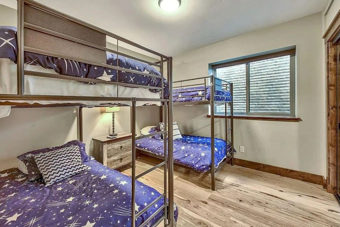 Une chambre de dortoir avec des lits superposés, une fenêtre avec des stores, une table de chevet avec une lampe et un plancher en bois.