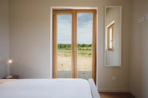 Un dormitorio con cama, mesita de noche, lámpara, ventana con vistas a un campo y espejo en la pared.