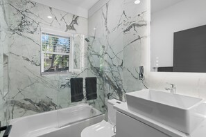 Ein modernes Badezimmer mit Badewanne, Toilette und Waschbecken, Marmorwänden und einem Fenster mit Jalousien.