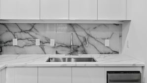 Una cucina moderna con mobili bianchi, una parete rivestita in marmo, un lavello in acciaio inossidabile e una lavastoviglie integrata.