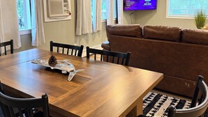 A dining area with a wooden table, chairs, a brown sofa, a TV, and a window with curtains.