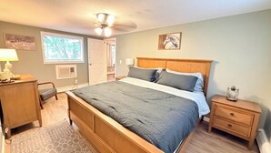 A bedroom with a bed, bedside tables, a desk, a chair, a window with blinds, a ceiling fan, and a door.
