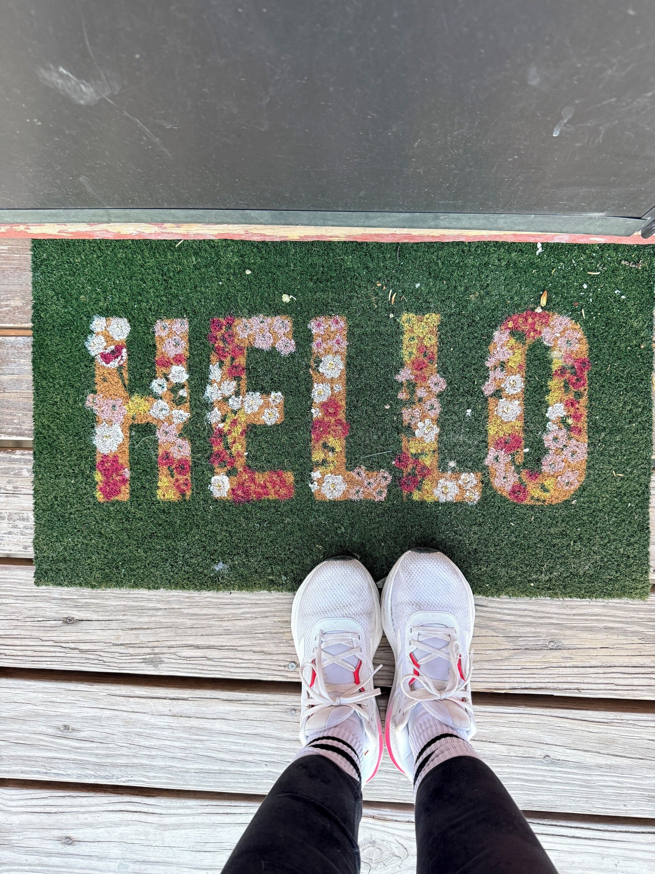 A welcome mat with 'HELLO' spelled out in colorful flowers, a wooden floor, and a pair of white sneakers.
