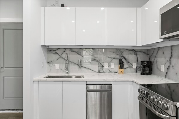 Cocina moderna con armarios blancos, salpicadero de mármol, electrodomésticos de acero inoxidable y horno empotrado.