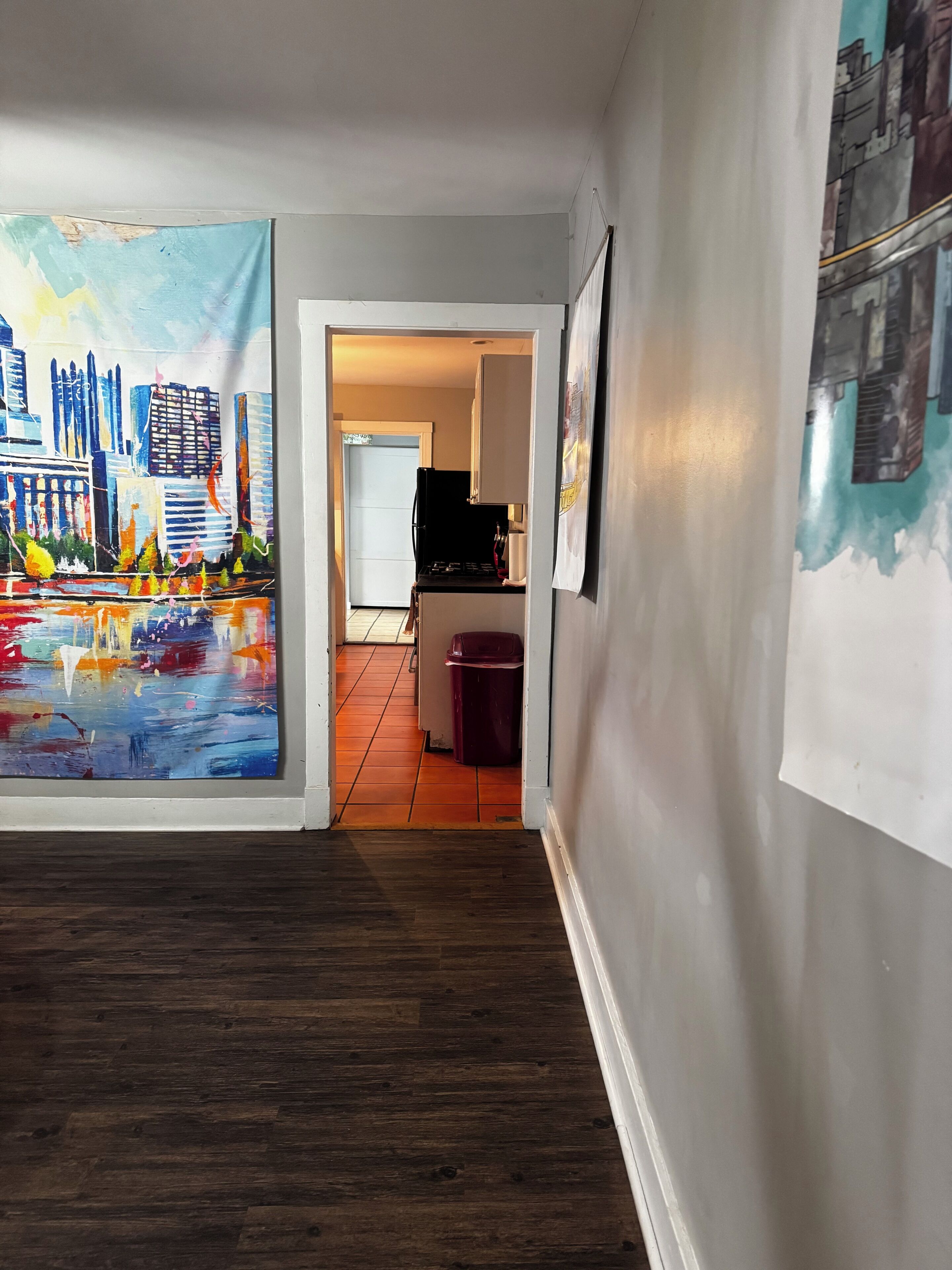 A hallway with a cityscape painting on the left, a kitchen area in the background, and a trash bin.