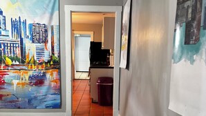A hallway with a cityscape painting on the left, a kitchen area in the background, and a trash bin. - 3-bedroom house in Southside Flats neighborhood of Pittsburgh. (Pittsburgh)