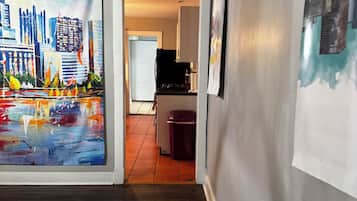 A hallway with a cityscape painting on the left, a kitchen area in the background, and a trash bin.