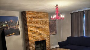A living room with a brick fireplace, a round coffee table, a dark sofa, a television, and a cityscape painting. - 3-bedroom house in Southside Flats neighborhood of Pittsburgh. (Pittsburgh)