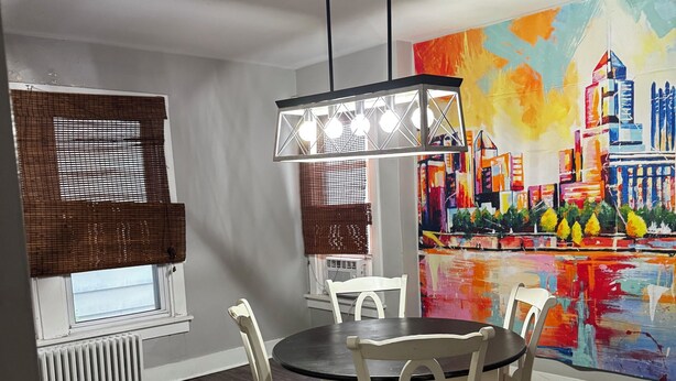 A dining area with a round table, white chairs, a hanging light fixture, a radiator, and a colorful cityscape painting on the wall. - 3-bedroom house in Southside Flats neighborhood of Pittsburgh. (Pittsburgh)