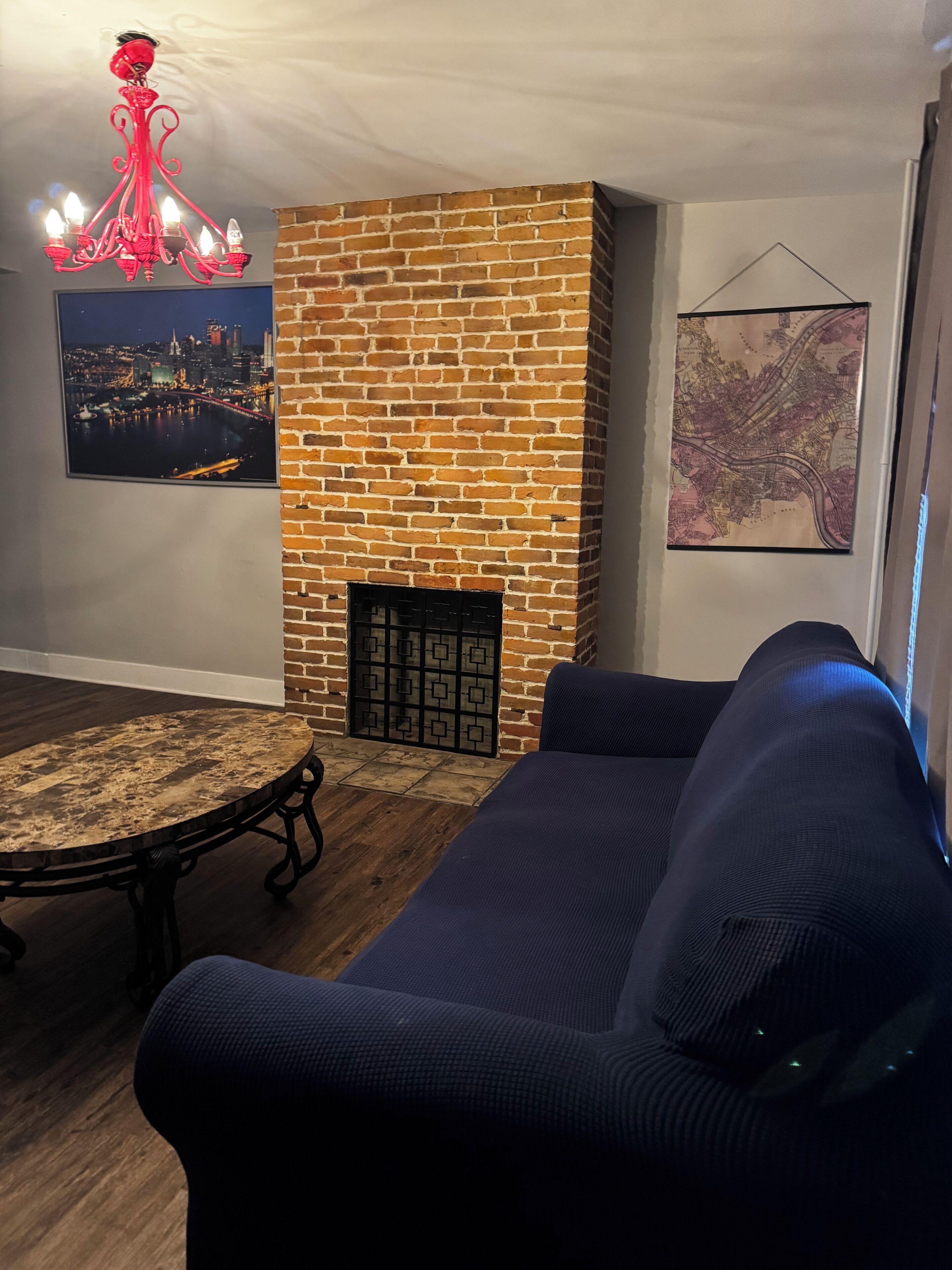 A living room with a brick fireplace, a round table, a blue sofa, a red chandelier, and two framed pictures on the walls.