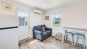 A compact kitchen with a white refrigerator, a grey sofa with a colorful pillow, and a window with a view of a building. - Charming Port Aransas Cottage Near Beach Access (Port Aransas)