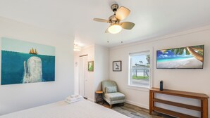 A bedroom with a bed, a ceiling fan, a painting of a ship, a chair, a TV, and a window with a view. - Charming Port Aransas Cottage Near Beach Access (Port Aransas)