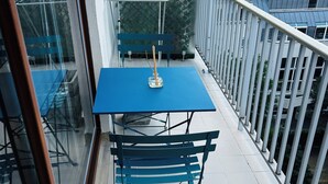 Un balcón con una mesa azul y sillas a juego, una pequeña mesa cuadrada con una vela, y una cerca de metal.