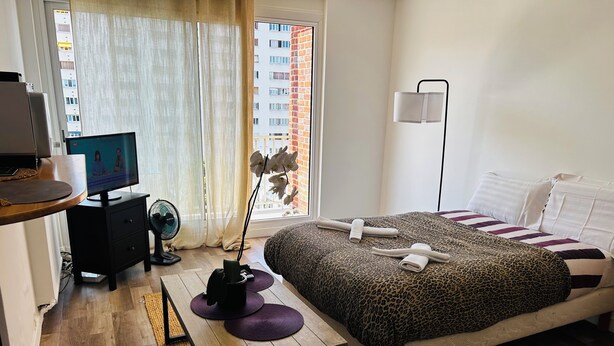 Un dormitorio con cama, escritorio con computadora, ventilador, ventana con cortinas y vista a un edificio.