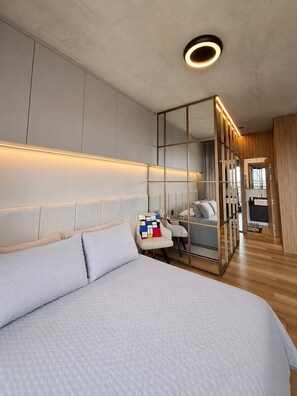 Ein modernes Schlafzimmer mit einem großen Bett, einer Sitzecke mit Sofa und Sesseln, einem raumhohen Spiegelkleiderschrank und einer kleinen Küchenzeile.