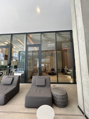 Ein moderner Innenbereich mit einer Lounge-Ecke, zwei Lounge-Stühlen und einem runden Hocker. Eine Glastür führt in einen Essbereich, und im Hintergrund ist eine Waschküche erkennbar.