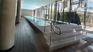 A modern indoor swimming pool with clear glass walls, wooden decking, and a view of the surrounding landscape. - Millenium Luxury Cozy Studio in Vila Ol Mpia (São Paulo)