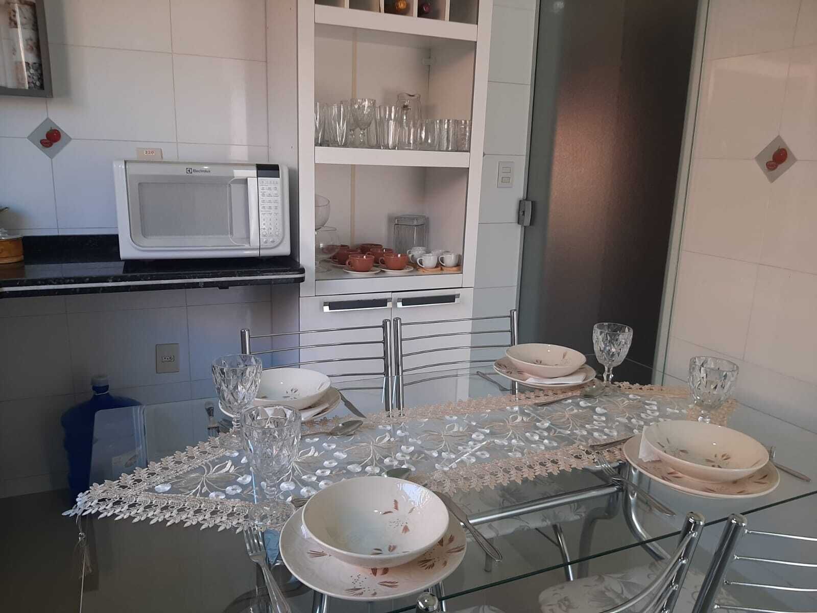 Un comedor con una mesa de vidrio dispuesta para cuatro personas, un microondas blanco y estantes con cristalería y vajilla.
