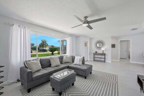A living room with a grey sectional sofa, a coffee table, a ceiling fan, and a view of a golf course through large windows. - Modern vacation home on the canal with a saltwater pool and private dock in the yacht club area (Cape Coral)