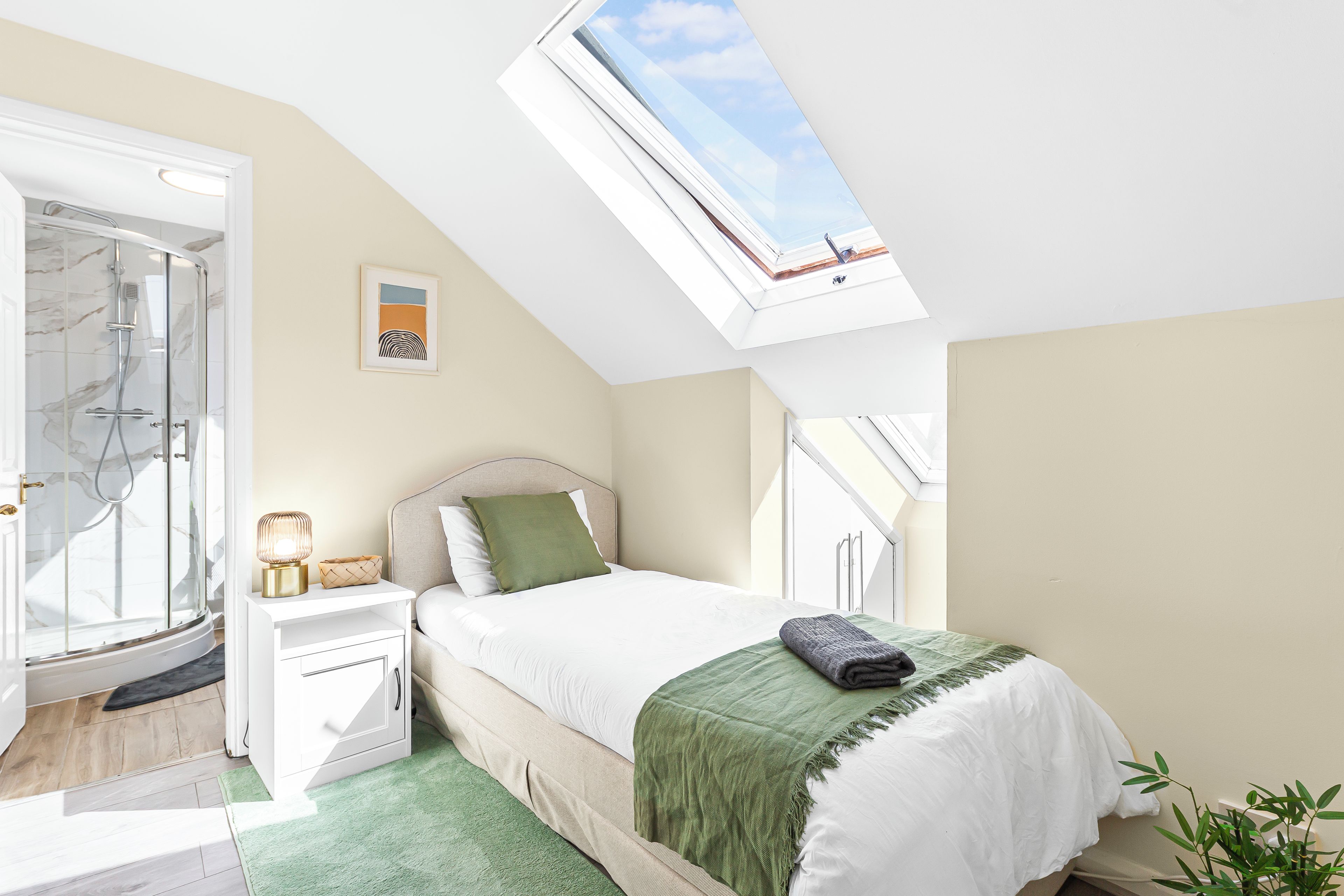 A bedroom with a skylight, a shower, a bedside table with a lamp, and a bed with a green throw.