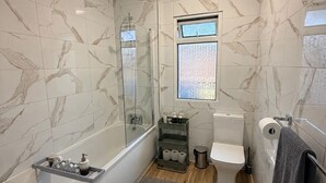 A bathroom with a bathtub, toilet, and shower area, marble walls, and a window.