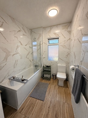 A bathroom with a bathtub, toilet, and shower area, marble walls, and a window.