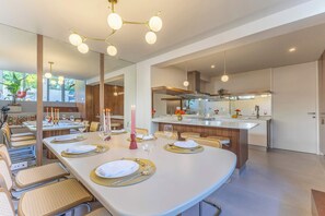 Un comedor moderno con una mesa larga dispuesta para una comida, una cocina con isla central y un espejo grande que refleja el espacio.